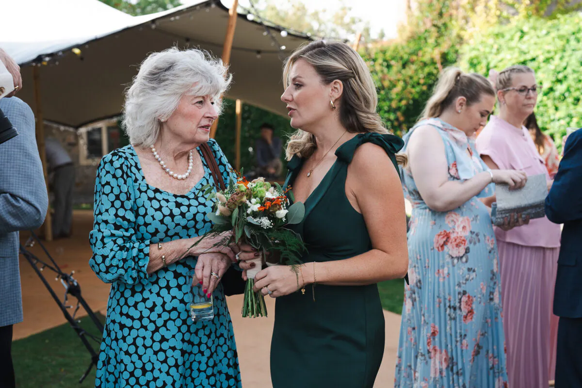 Two wedding guests chat together outdoors near a marquee, with one woman holding a small hand-tied floral bouquet.