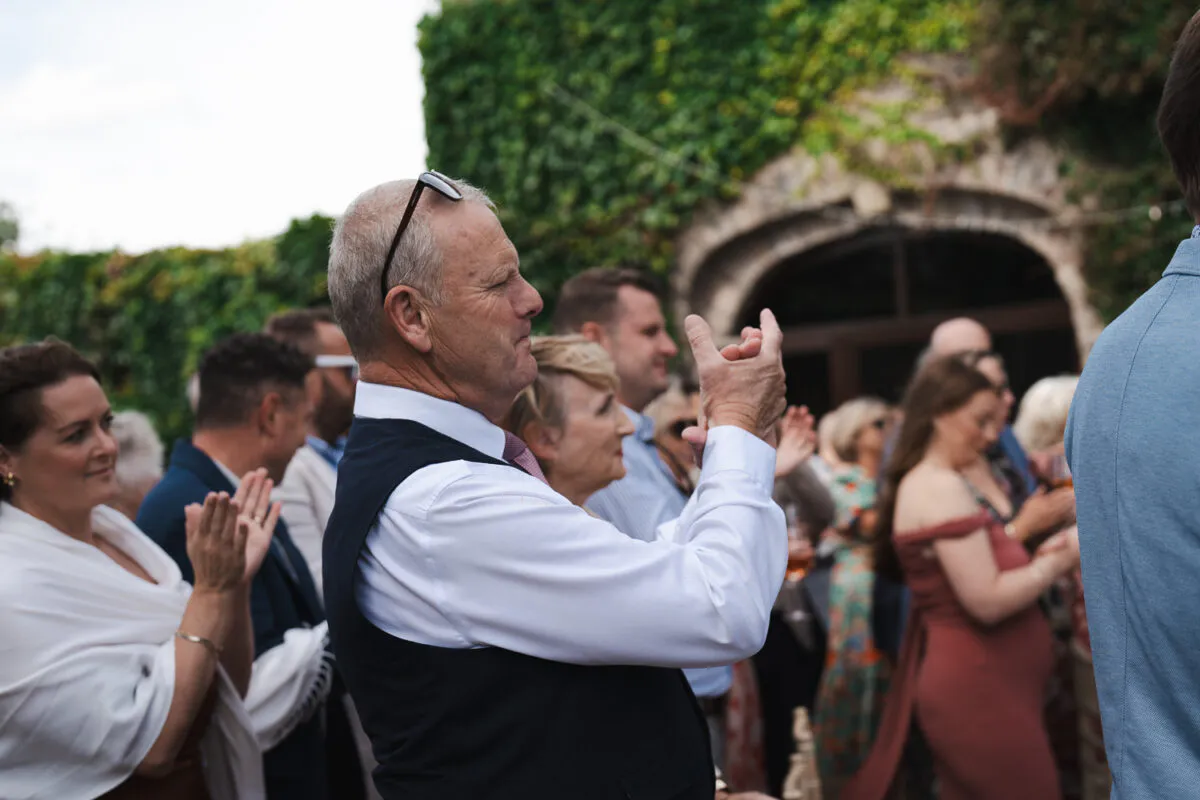 Wedding guests clap and cheer outdoors in front of a stone archway covered in ivy.