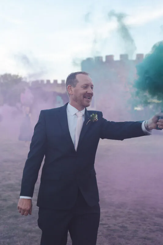 A smiling groom in a navy suit holding a smoking flare in front of an ancient castle ruin.