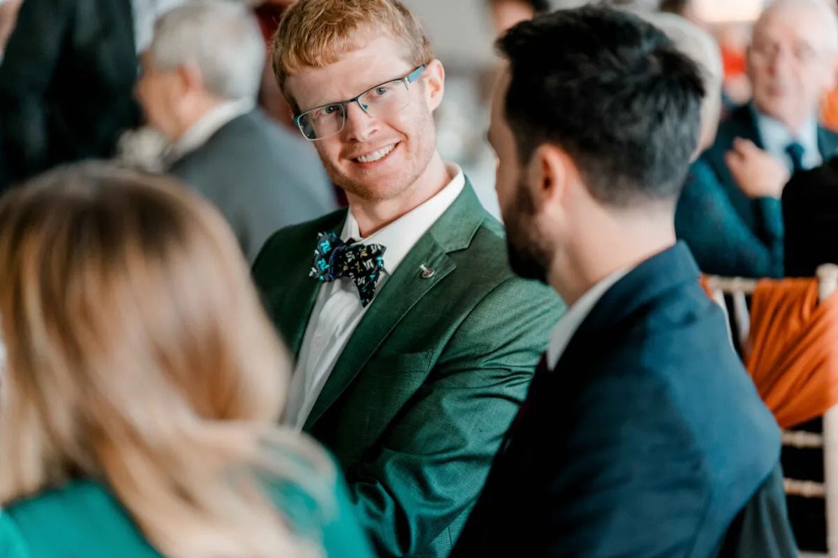 A smiling guest wearing a green suit and patterned bow tie chats and mingles with other wedding attendees during the drinks reception.