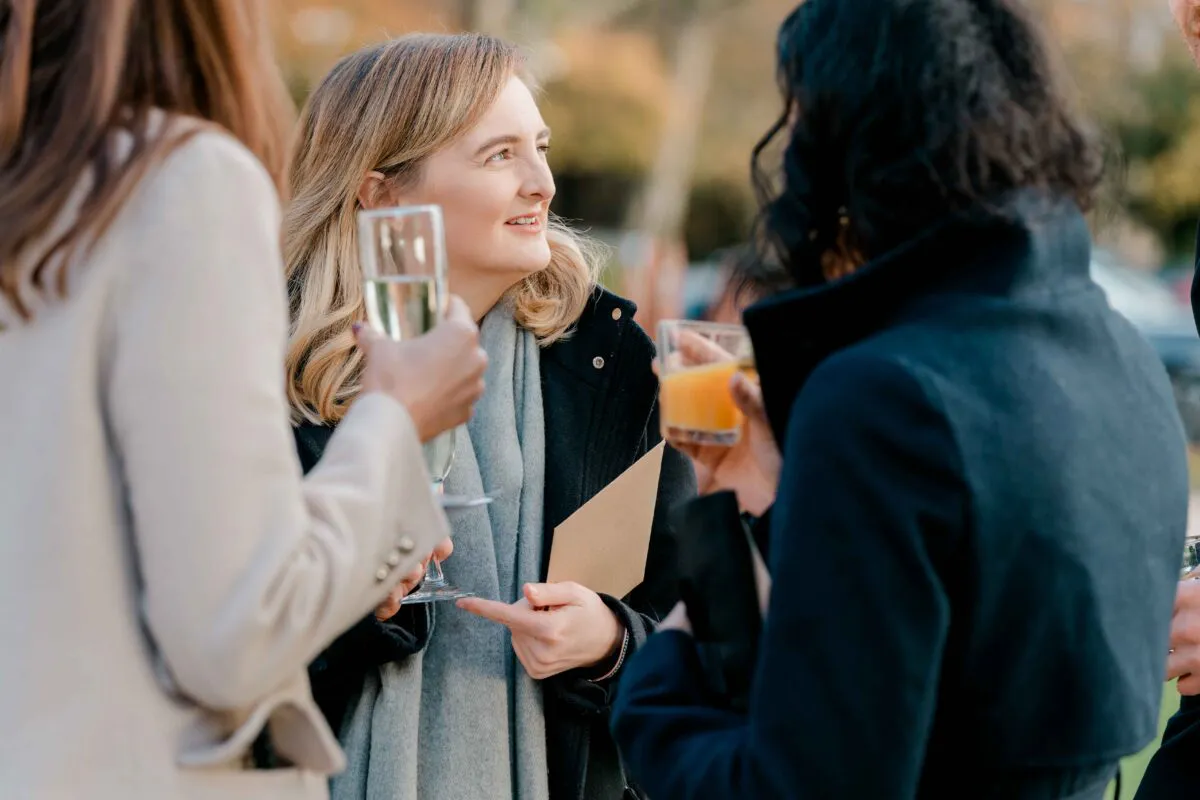 Wedding guests mingle outdoors during the drinks reception, holding drinks and chatting happily in natural daylight.
