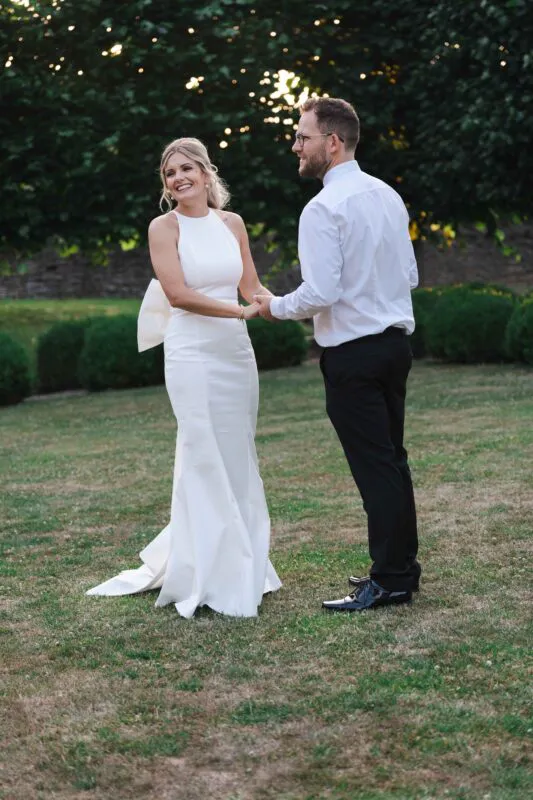A bride in a high-necked crepe gown with a large bow detail and a groom in shirt sleeves hold hands and laugh together on a grassy lawn during golden hour.