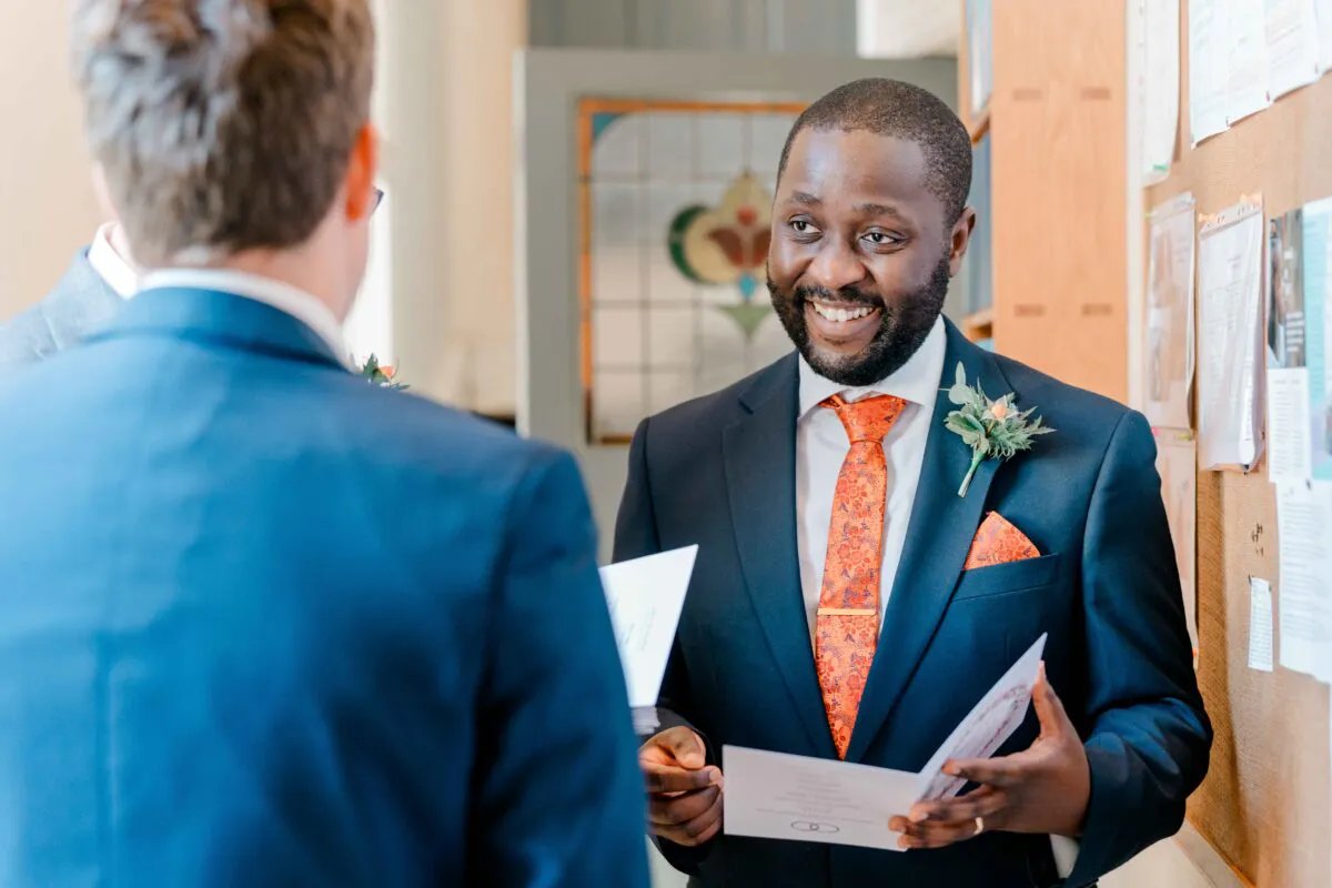A smiling groom wearing a dark suit and orange patterned tie holds an order of service while chatting with another guest during the wedding day.