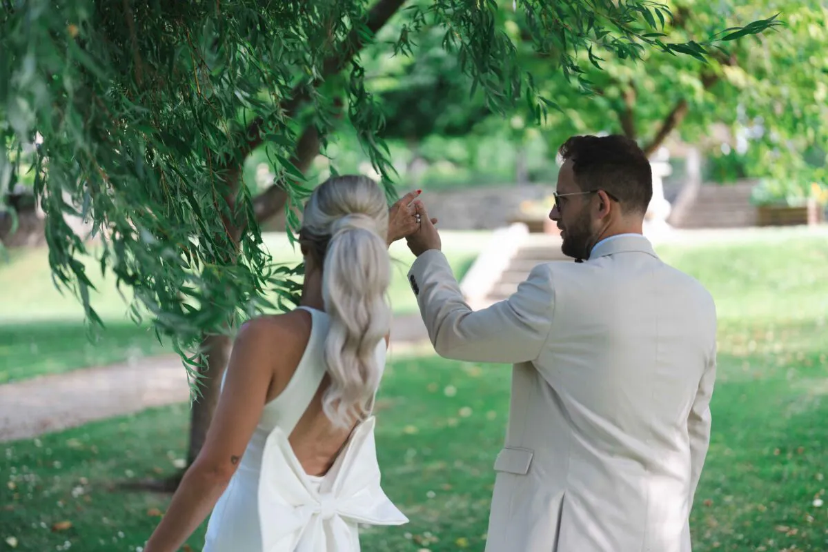 A newlywed couple walks hand in hand through a lush green garden, beautifully framed by the drooping branches of a weeping willow tree.