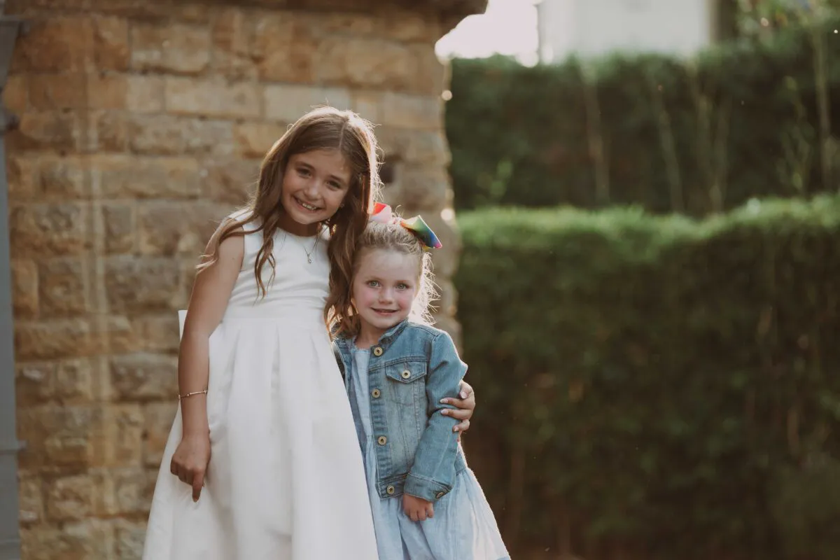 Two young girls smile happily while embracing outdoors near a rustic stone wall during a wedding celebration.
