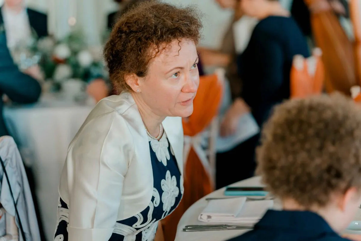 A guest with curly brown hair wearing a patterned dress and white bolero is seated at a wedding breakfast table, engaged in conversation.