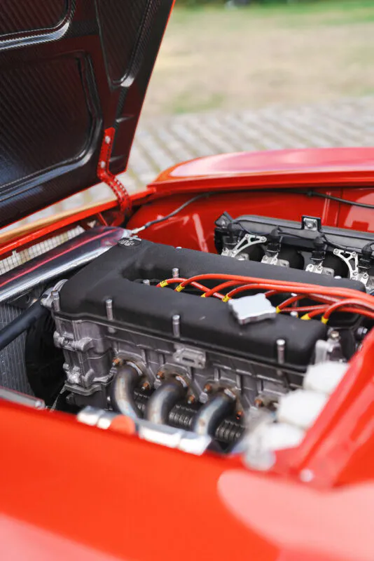 A close-up view of a vibrant red vintage sports car with its bonnet open, revealing the polished engine and mechanical details.