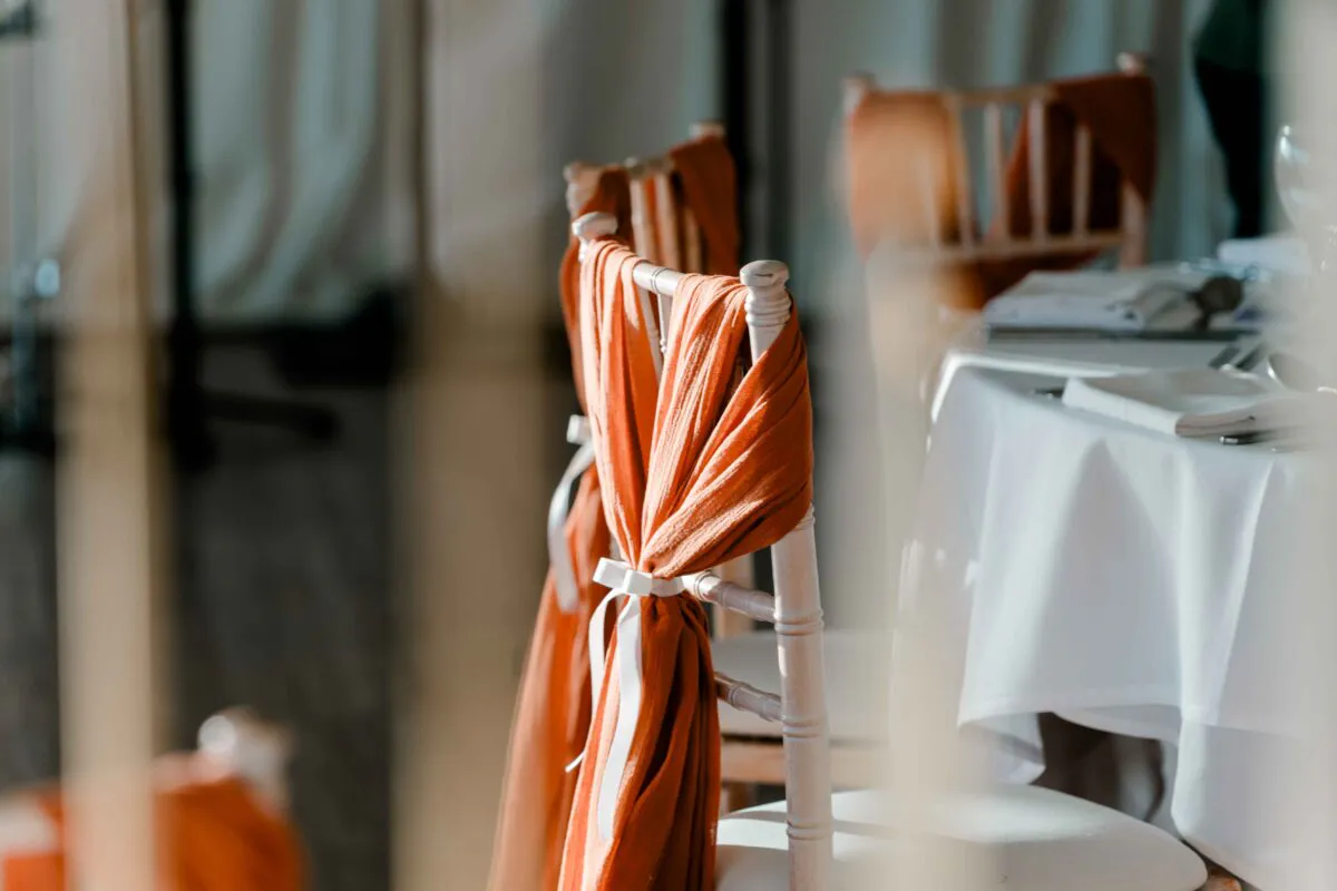 Chairs draped with flowing terracotta fabric and white ribbon sit beside a neatly set wedding table with white linens.