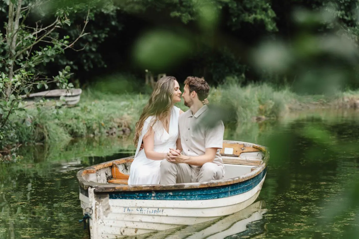 A bride and groom share a tender moment, holding hands and leaning in for a kiss while seated inside a wooden rowing boat on a calm lake.
