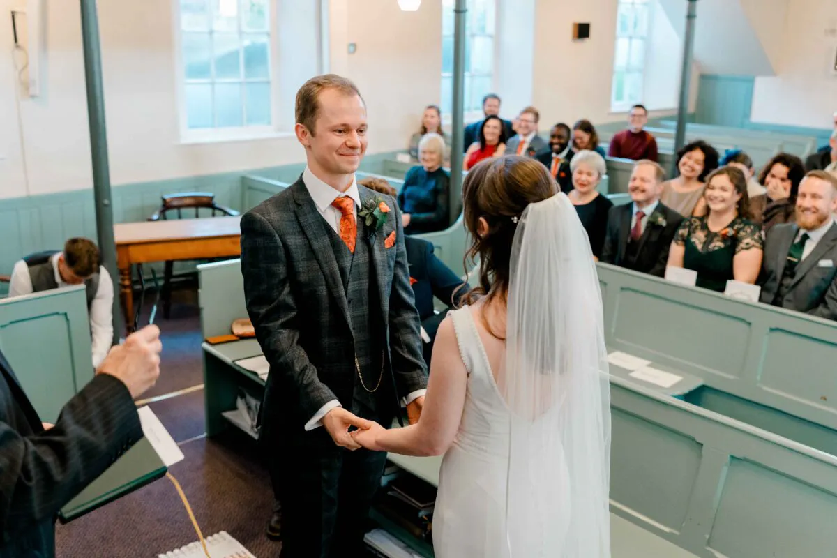 A smiling groom in a tweed suit holds hands with his bride during an indoor chapel wedding ceremony while guests watch from the pews.