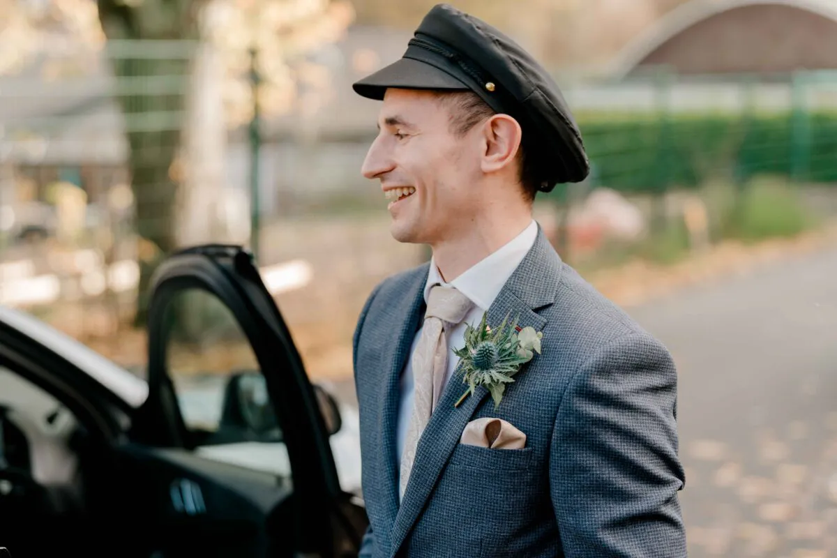 A smiling groom wearing a grey patterned suit, tie, and cap stands beside an open car door with a thistle buttonhole.