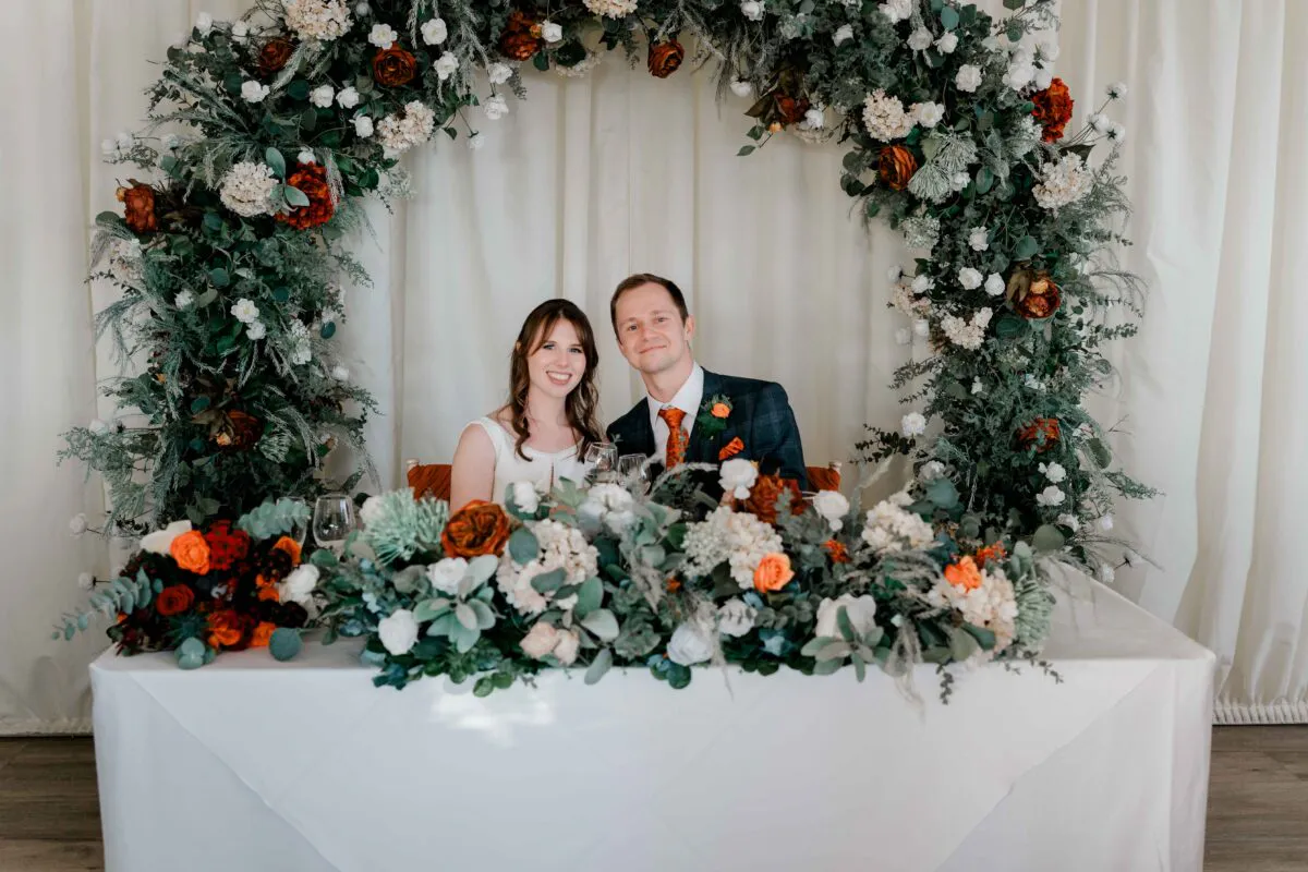 A newlywed bride and groom sit smiling behind a heavily decorated sweetheart table adorned with lush white and burnt orange floral arrangements beneath a matching circular arch.