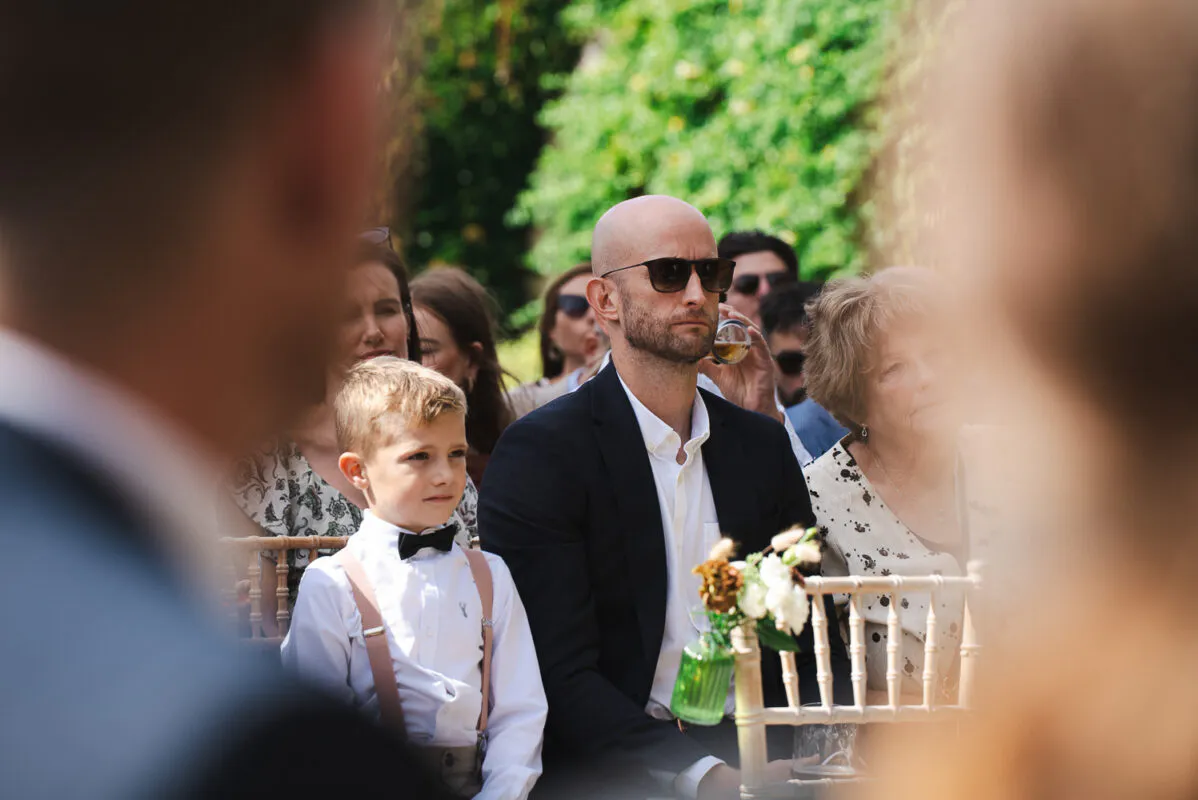 A young page boy in a white shirt and braces sits beside a smartly dressed man wearing sunglasses among wedding guests attending an outdoor ceremony.