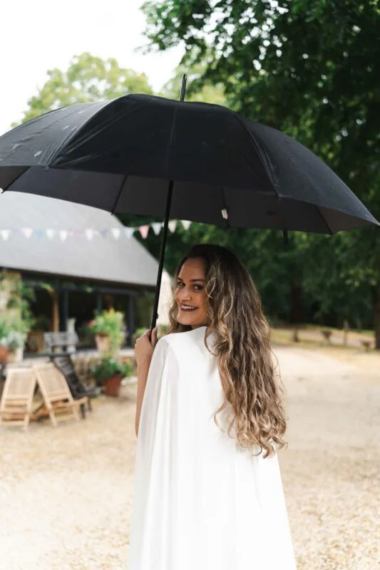 A smiling bride with long curly hair looks over her shoulder while holding a black umbrella outdoors near a rustic building decorated with bunting.