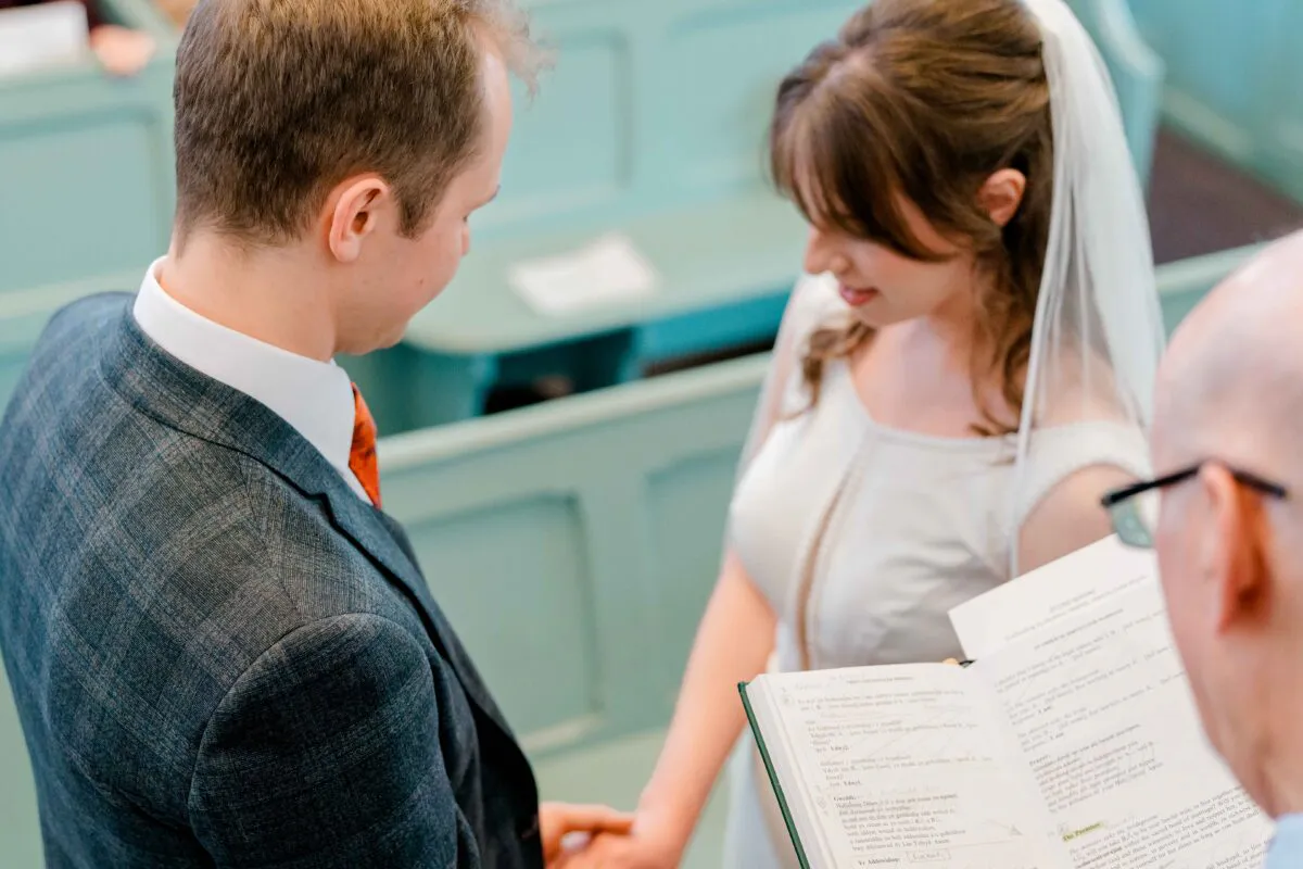 A bride and groom exchange vows inside a traditional church with wooden pews while the officiant holds the service book.