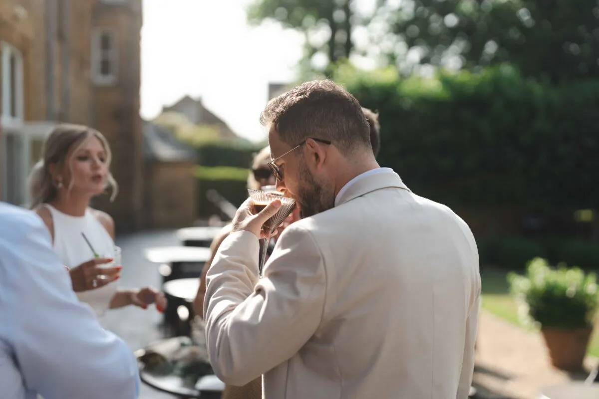 A groom in a stone linen suit enjoys an espresso martini from a coupe glass during an outdoor drinks reception in the sunshine.