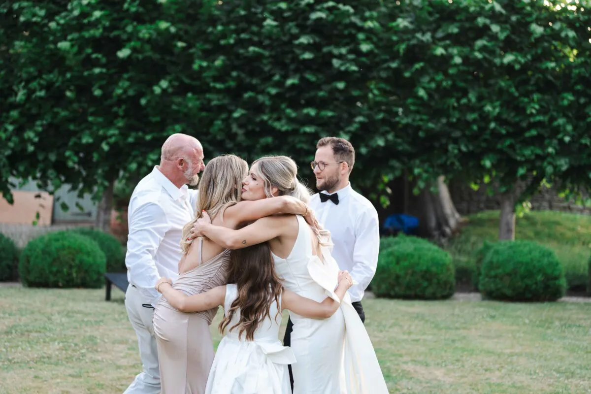 A bride in a white gown and a small flower girl embrace lovingly in a lush garden setting as family members look on smiling.