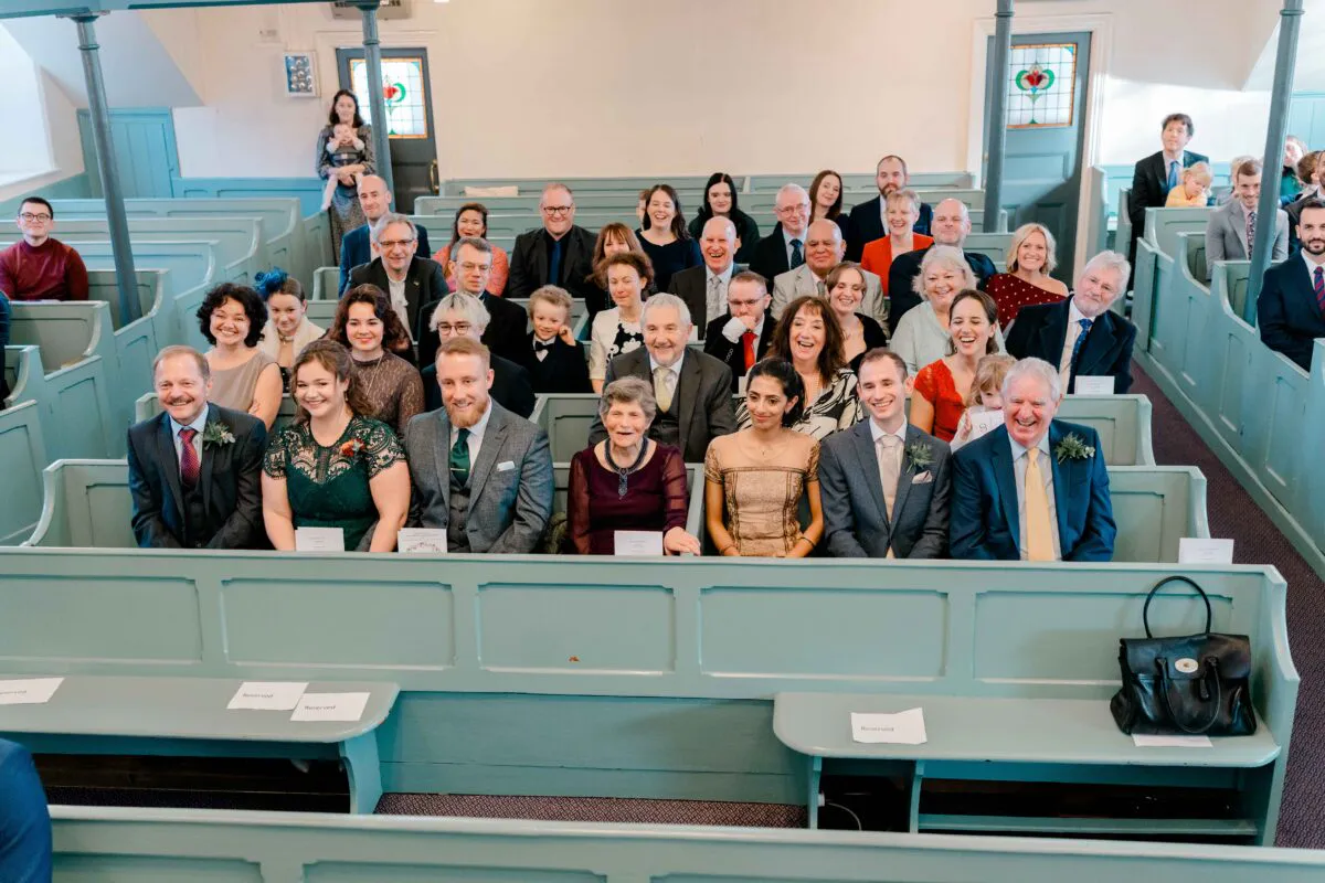 A large congregation of smiling and laughing wedding guests seated in wooden pews inside a historic church for the ceremony.