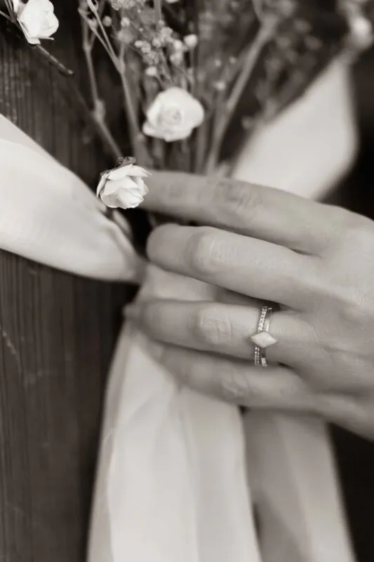 Close-up of a hand wearing stacked wedding rings while delicately holding a small bouquet of white roses tied with a ribbon.