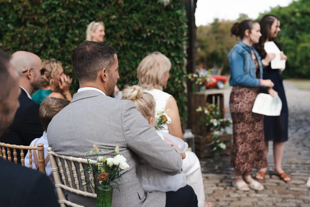 An over the shoulder shot of wedding guests seated on chivari chairs outdoors during a ceremony, with a man in a grey suit holding a young flower girl.