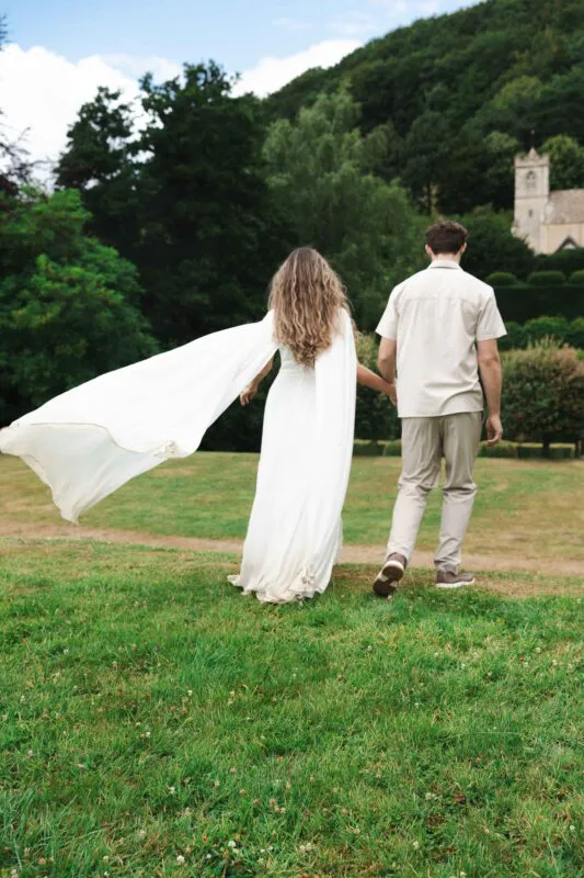 A newlywed couple walking hand in hand across a lush green lawn with a building and rolling hills in the background, featuring a dramatic flowing cape on the bride.