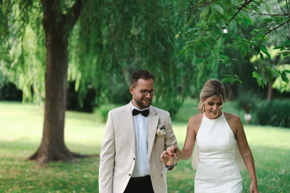 A newlywed couple happily walk hand in hand through a lush green woodland, framed by overhanging tree branches.