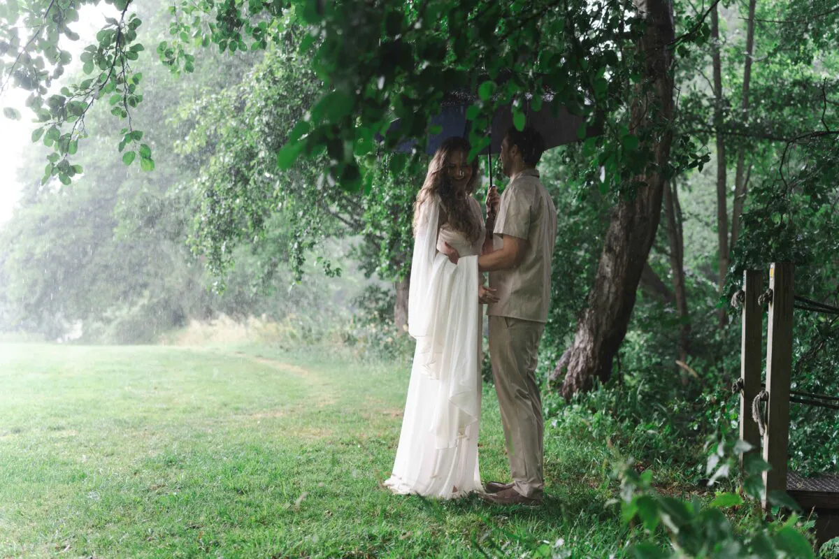 A newlywed couple shares a tender moment beneath an umbrella in a lush, rainy woodland setting.