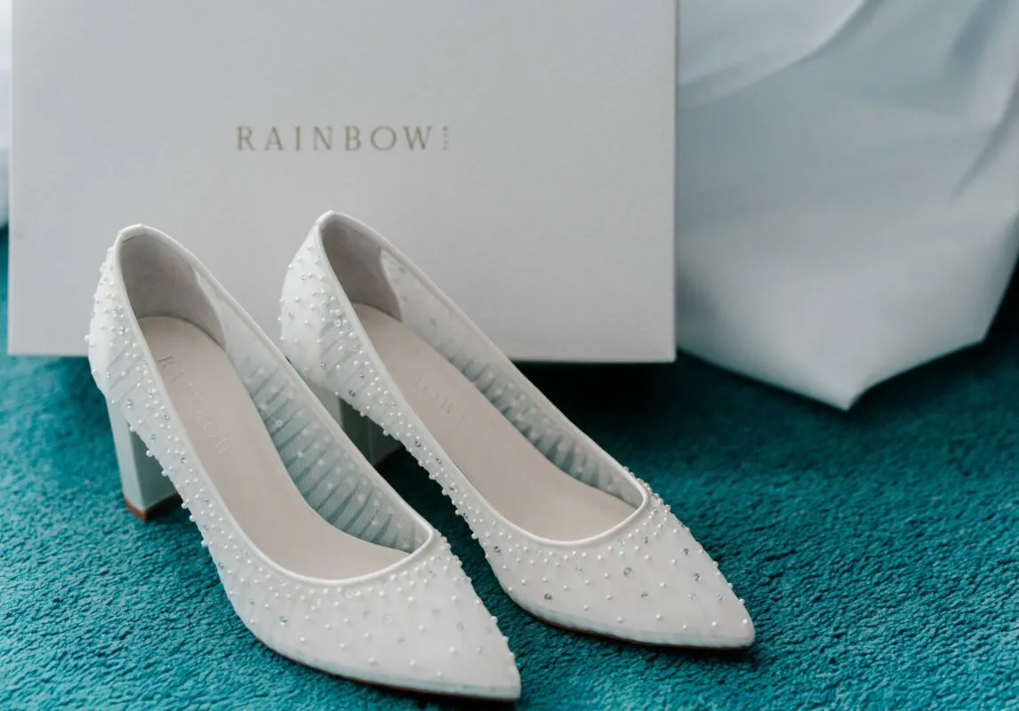 A pair of white block heel bridal shoes embellished with delicate pearls sit proudly in front of their branded storage box on a textured teal carpet.