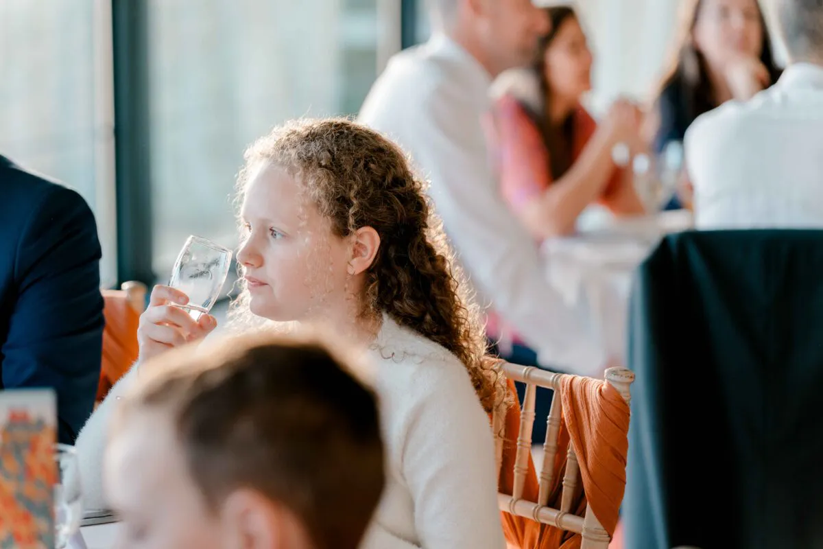 A young wedding guest with curly hair holds a glass of water at the reception table, illuminated by warm golden sunlight.