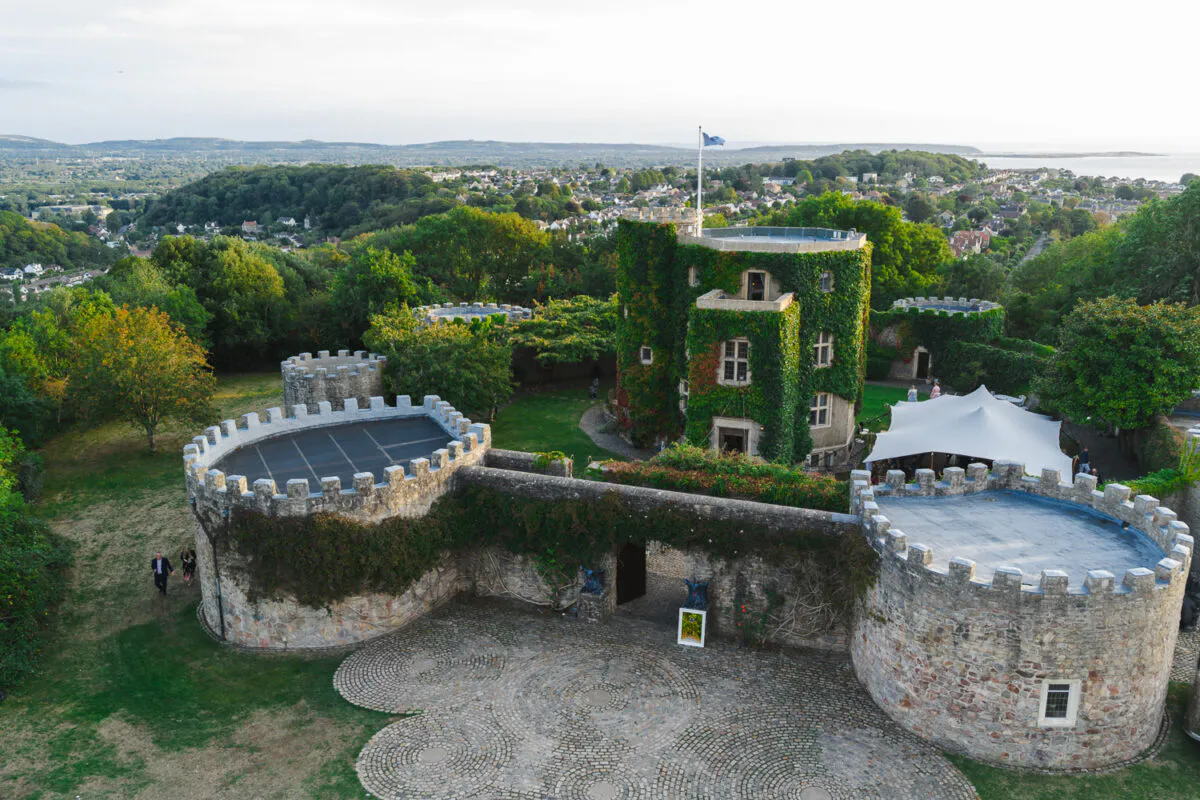 An aerial view captures a historic stone castle venue featuring ivy-covered towers, a cobbled courtyard, and a white stretch tent set amidst lush green surroundings.