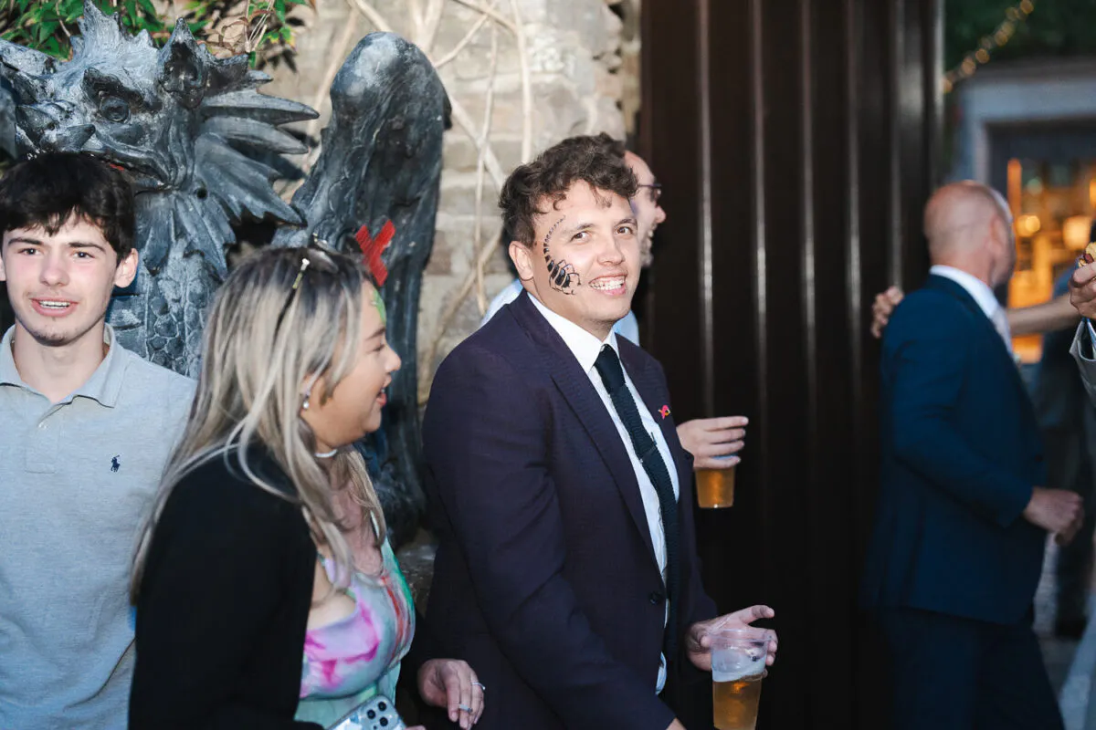 A smiling guest with face art on his cheek stands holding a drink outdoors among mingling wedding attendees near a stone gargoyle sculpture.