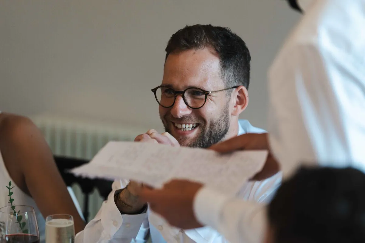 A smiling guest wearing glasses reacts with amusement during the wedding reception speeches while holding a speech paper.