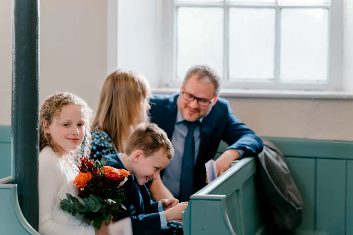 A young flower girl holding a small bouquet of orange and red roses smiles towards the camera while sitting in a church pew with other guests.