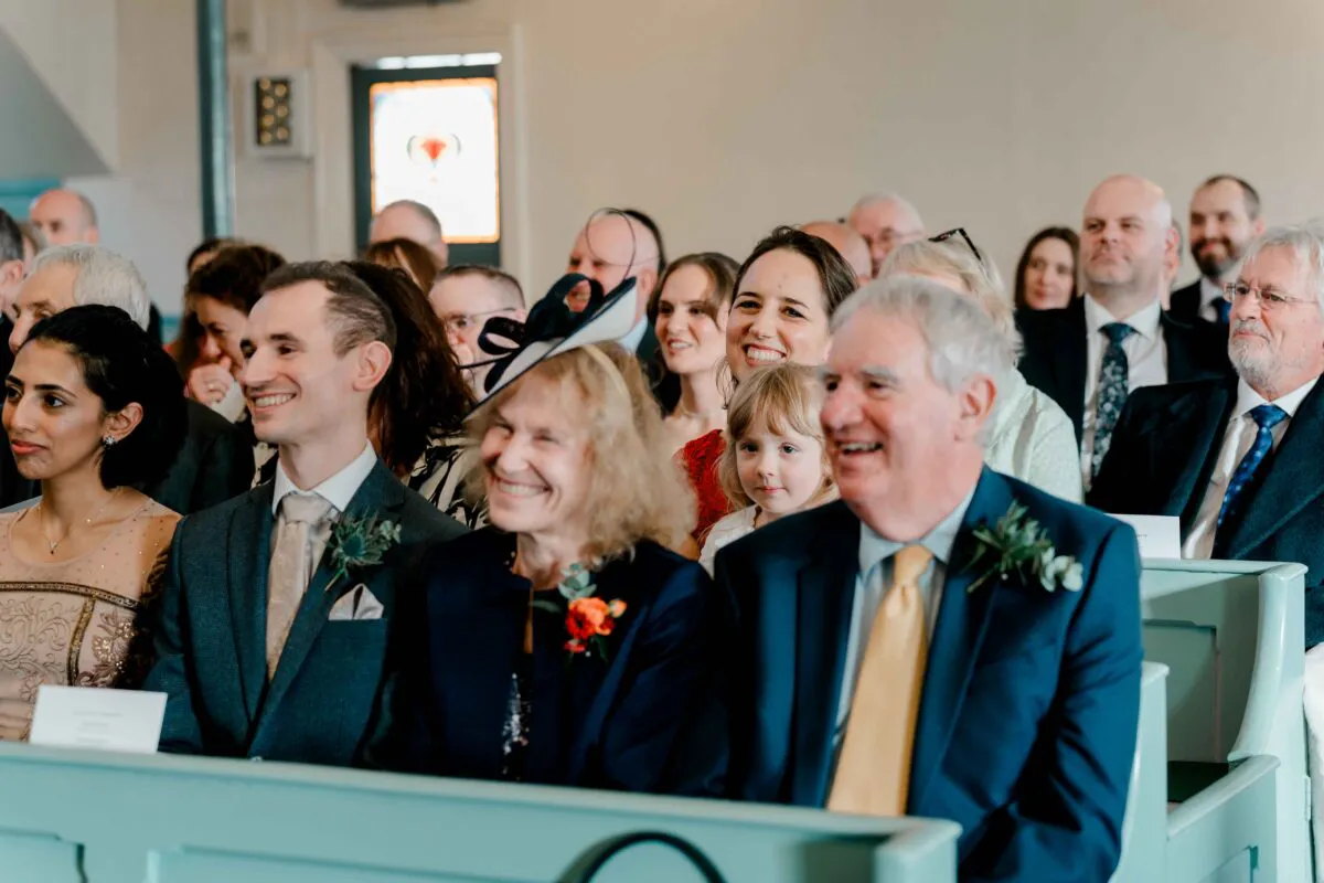 Happy wedding guests smiling and laughing together while seated in church pews during the marriage ceremony.