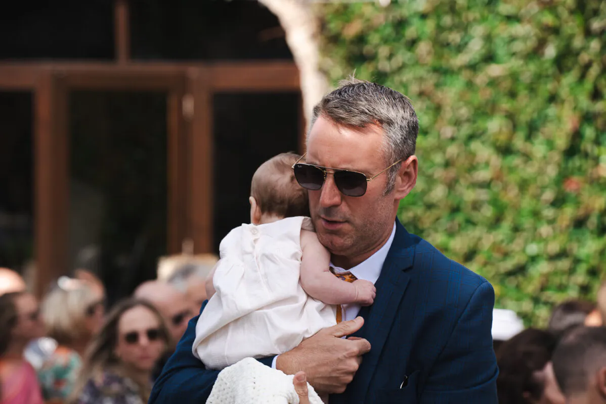 A guest wearing a navy patterned suit and sunglasses holds a baby dressed in white during an outdoor wedding celebration.