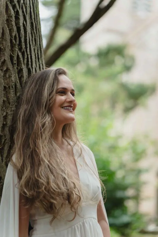 A smiling bride with long flowing hair stands leaning against an ancient oak tree in a woodland setting during her wedding portraits.