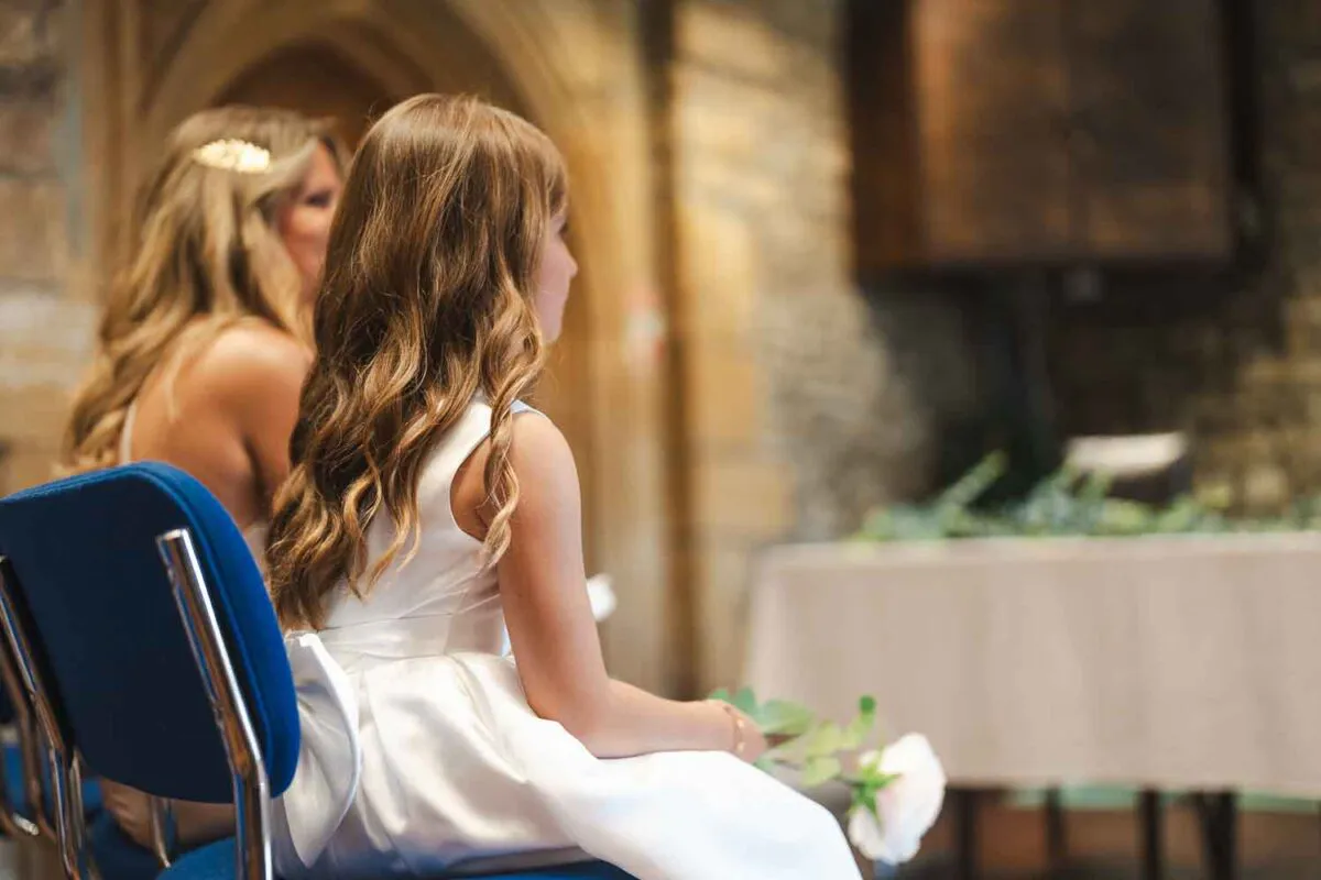 A young flower girl in a white dress sits attentively during a church wedding ceremony, viewed from behind in soft natural light.