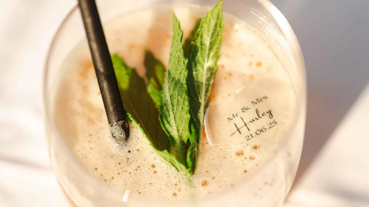 A close-up shot of a signature wedding cocktail garnished with fresh mint leaves and a custom stamped ice cube reading Mr & Mrs Hurley.