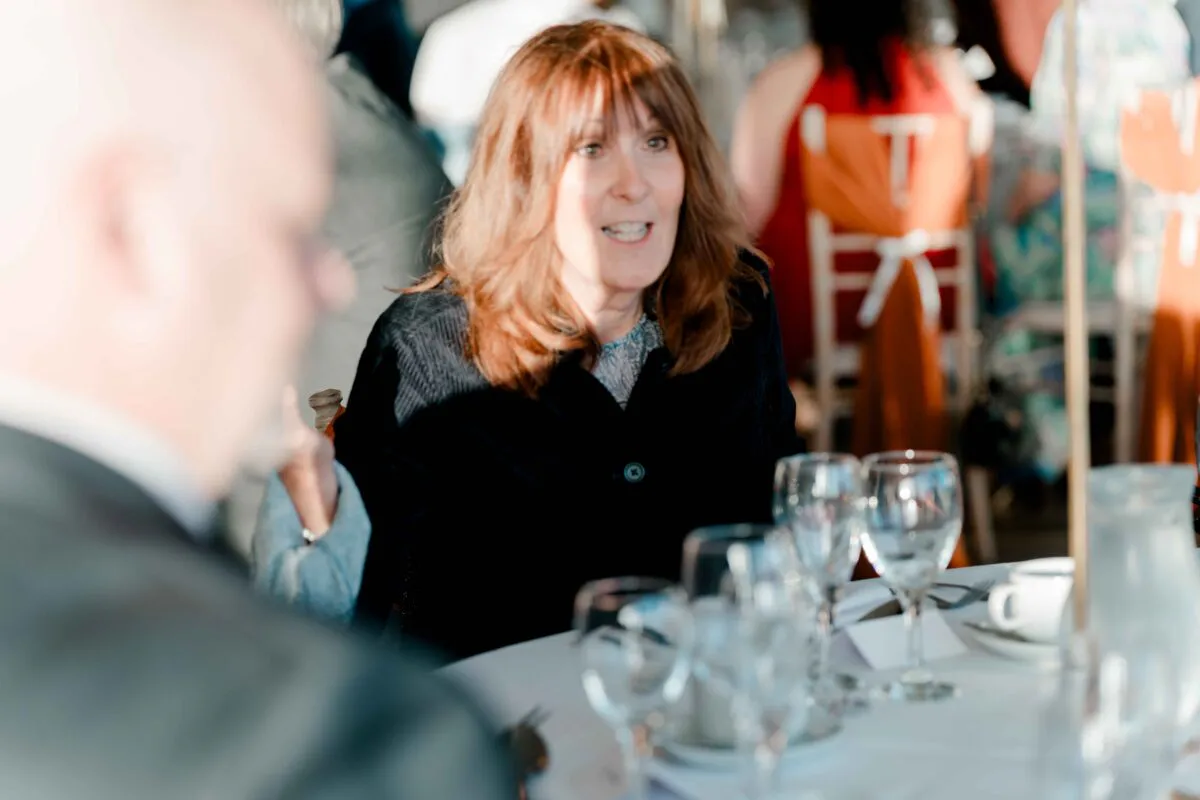A female wedding guest with auburn hair smiles and talks animatedly during the wedding breakfast seated at a round table.