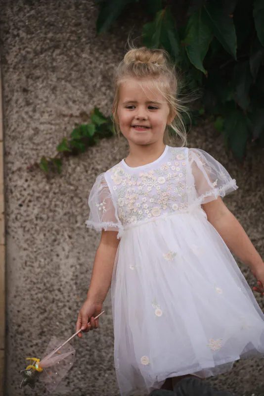 A young flower girl with her hair in a high bun wears a sheer embroidered dress while holding a small wand against an ivy covered stone wall.