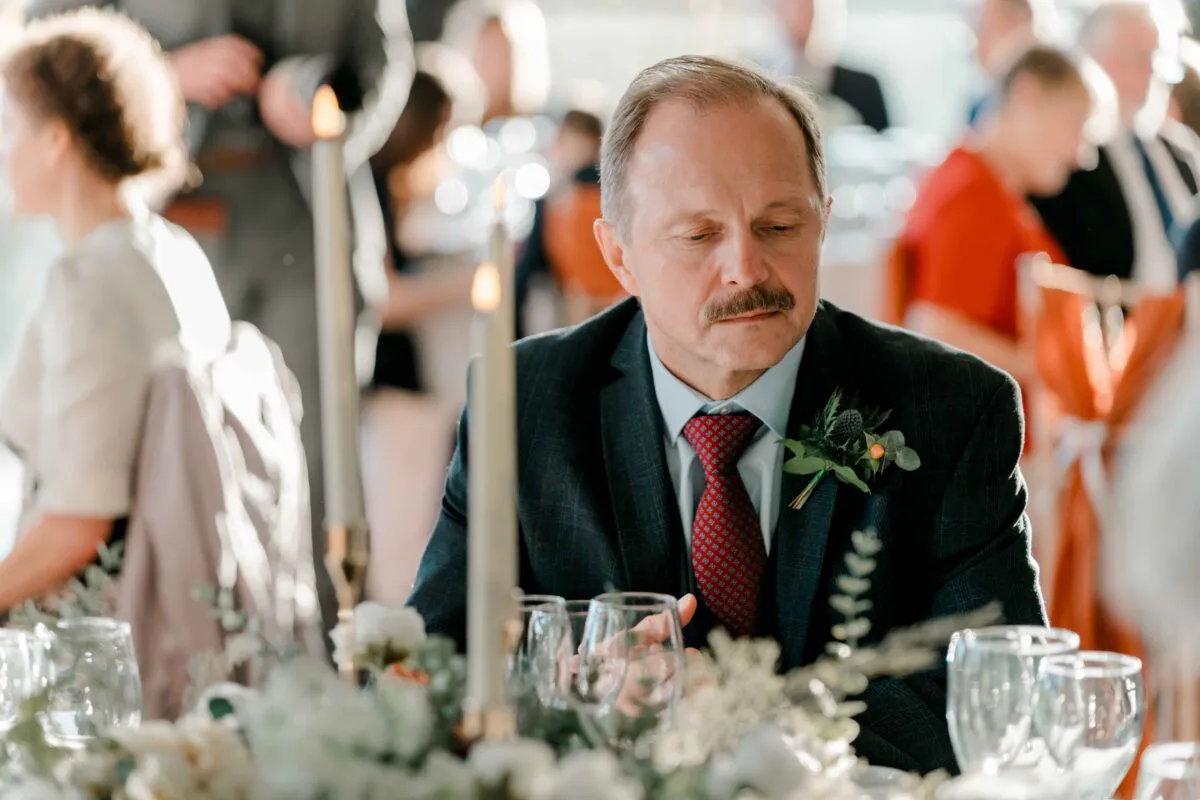 A guest in a dark suit and patterned tie sits attentively at a wedding reception table adorned with candles and floral centrepieces.