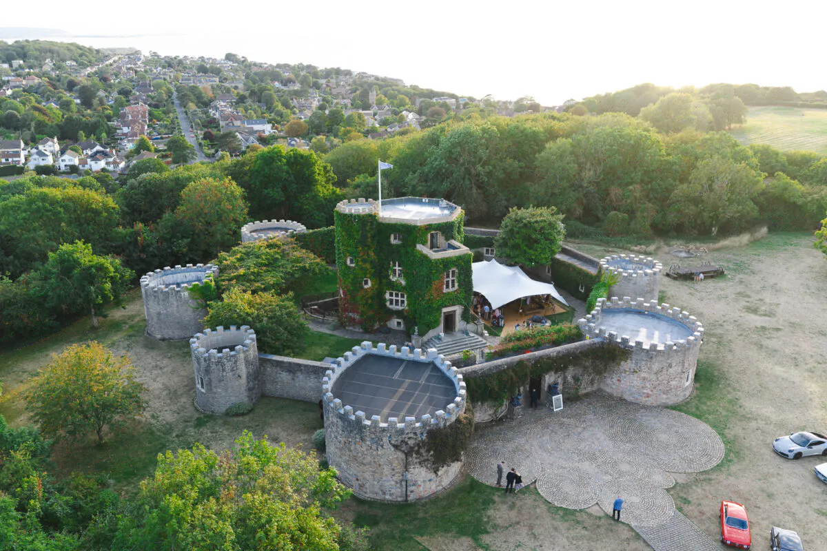 An aerial drone shot captures a historic stone castle venue surrounded by lush greenery, featuring a white stretch tent pitched in the courtyard and a cobbled driveway below.
