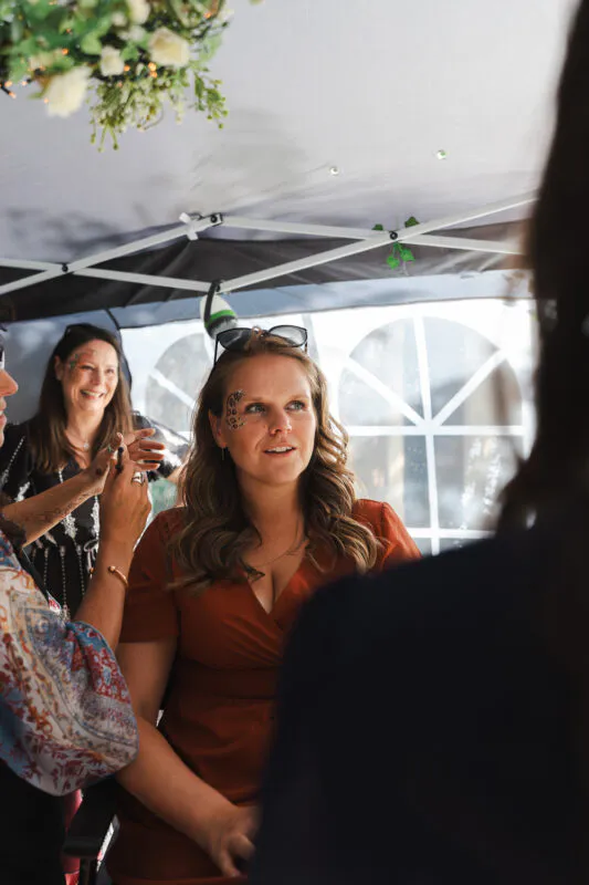 A guest sits smiling inside a marquee while having face art applied during a festival-styled wedding celebration.