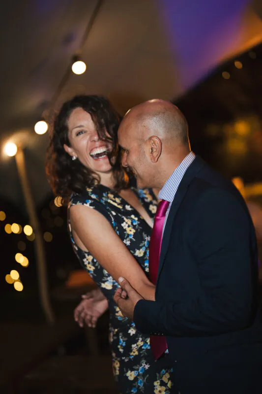 A smiling couple dances closely together at an evening reception, illuminated by warm string lights and ambient overhead lighting.