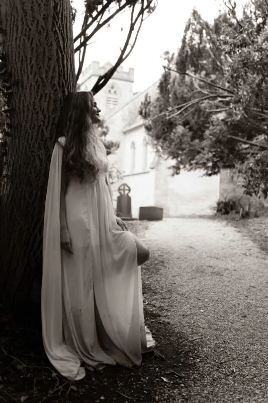 A solo bride wearing a flowing dress with a matching cape leans against a large tree in a churchyard in this sepia-toned portrait.