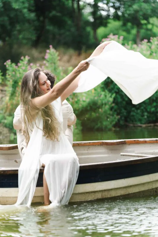 A newlywed couple laughing playfully while sitting in a wooden boat on a lake, with the bride holding up a white fabric.