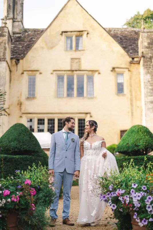 A bride and groom walk hand in hand through an English country garden path, framed by manicured topiary and a historic stone manor house.