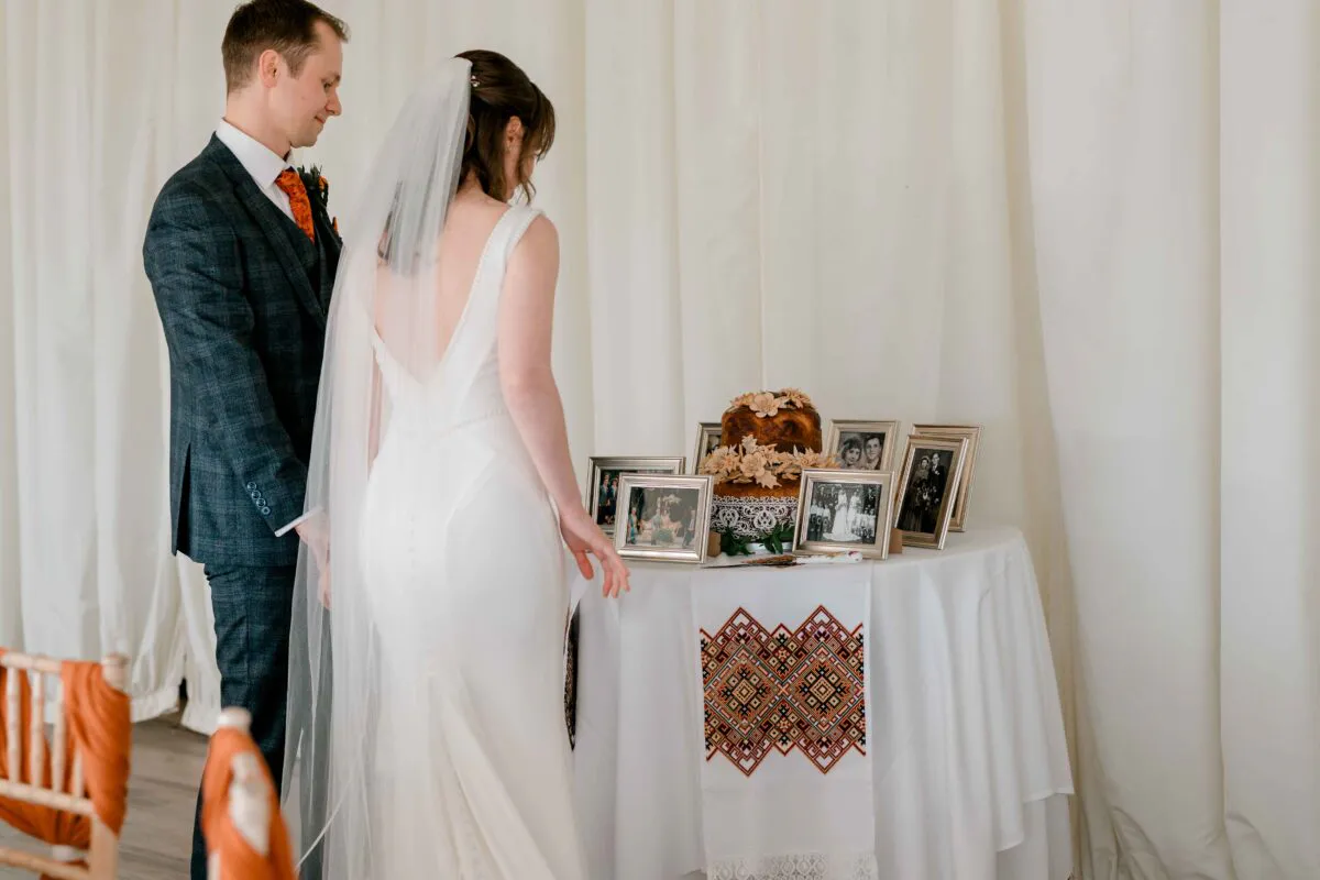A bride in a sleek crepe gown with a chapel veil and a groom in a checked suit look admiringly at a memory table adorned with framed family photographs and a decorative cloth.