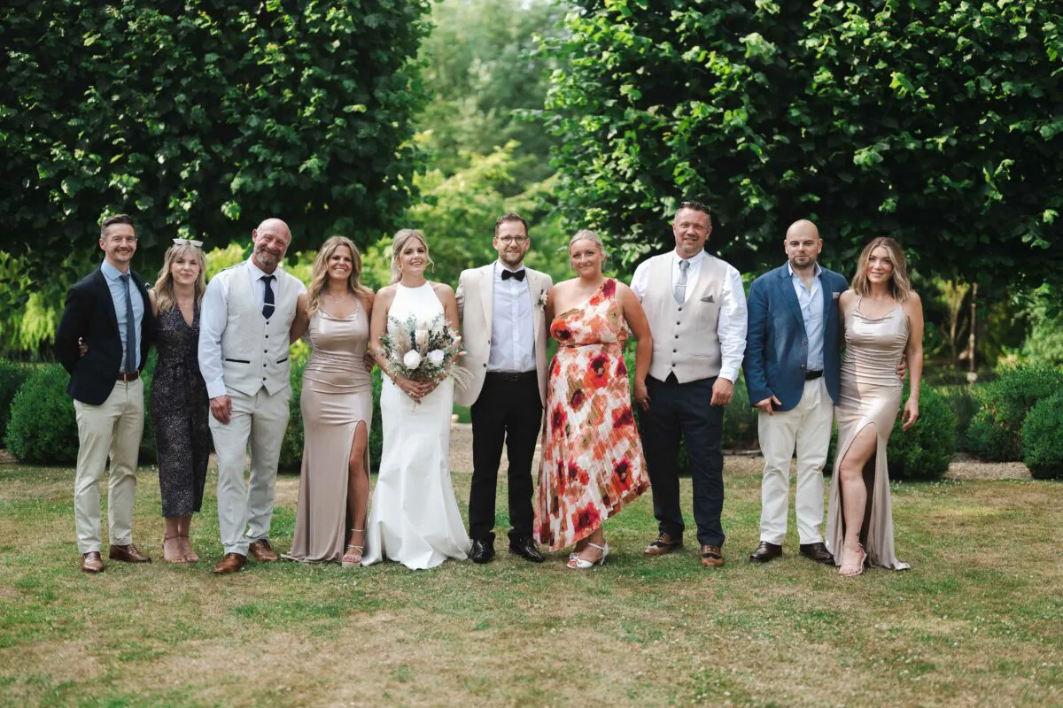 The bride and groom pose outdoors in a lush garden setting alongside their wedding party, featuring bridesmaids in champagne satin gowns and guests in smart attire.