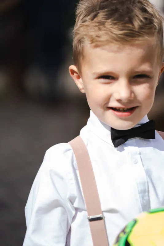 A young page boy wearing a white shirt, black bow tie, and pink braces smiles brightly outdoors in the sunshine.